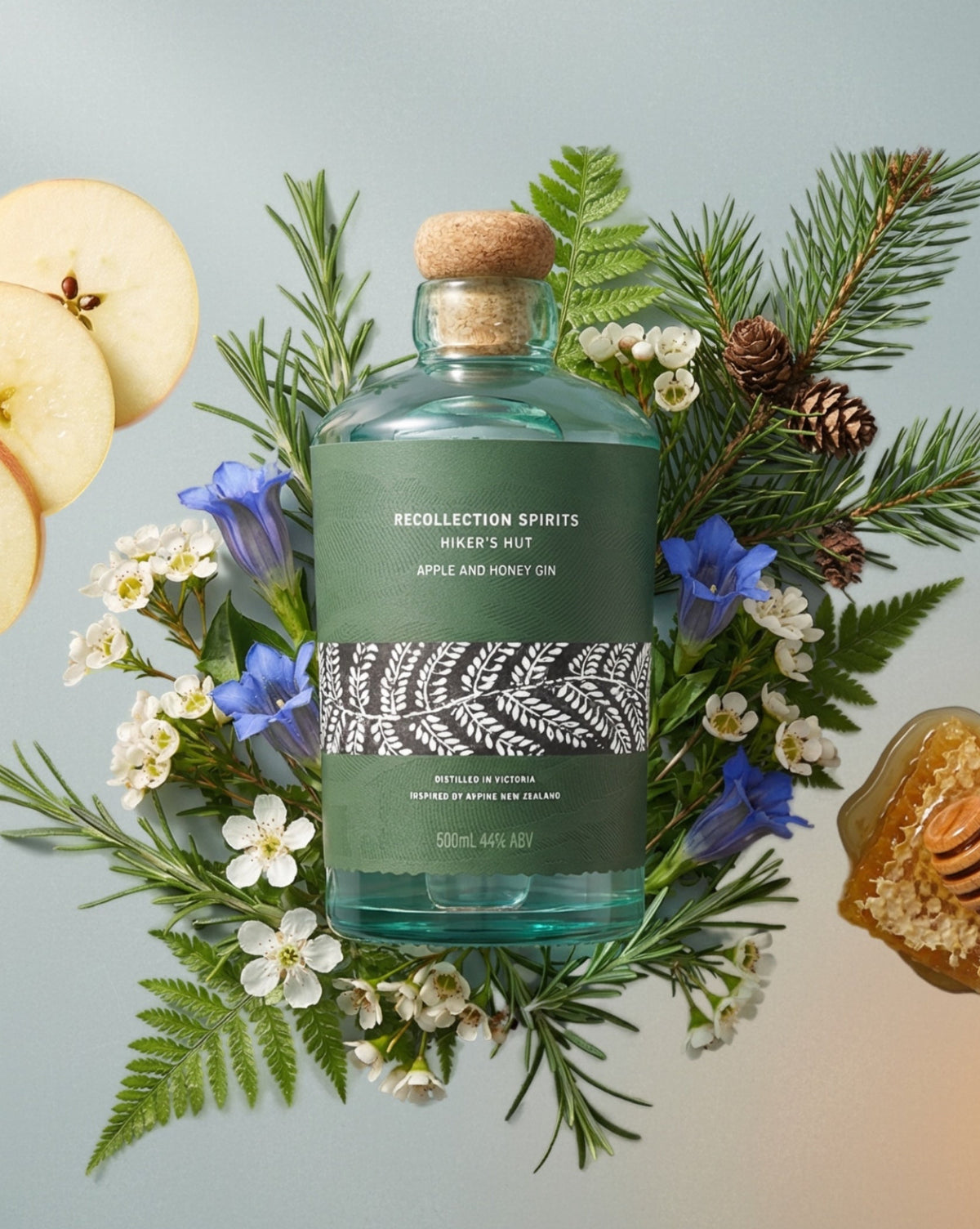 Bottle of Recollection Spirits Apple & Honey Gin surrounded by apples, flowers, and foliage on a light blue background.