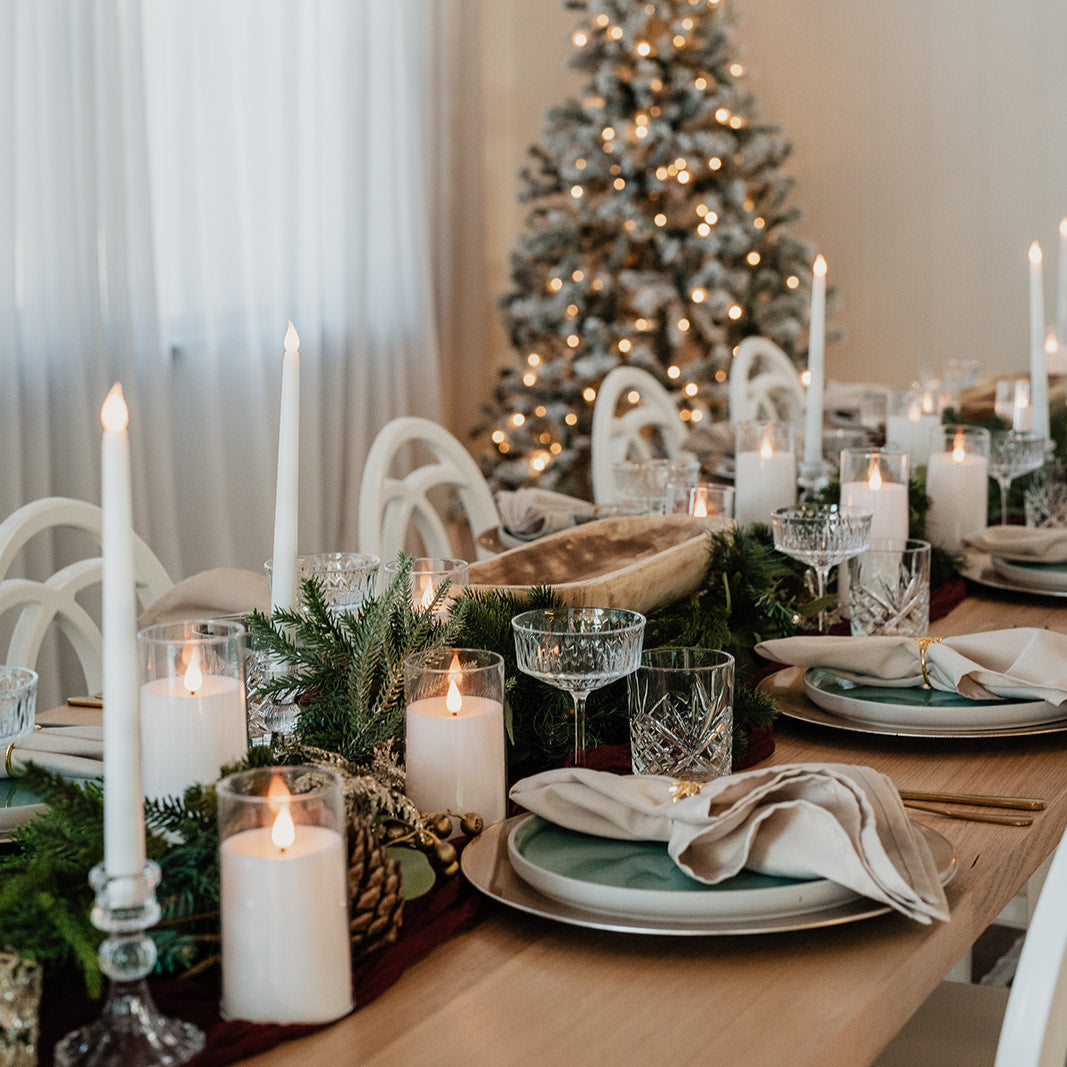 Dining table set for a Christmas meal with candles, glasses, and a decorated tree in the background.